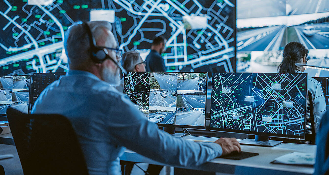 Control room with multiple screens showing traffic feeds and maps, focused middle-aged man in blue shirt with headset monitoring.