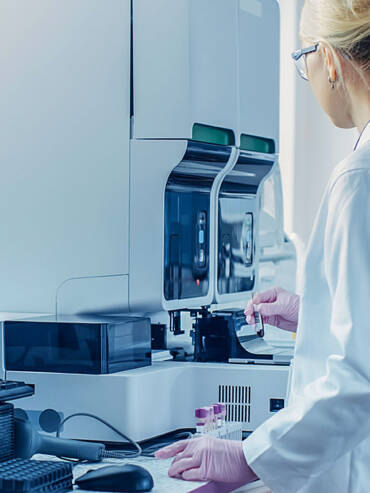 Female scientist in lab coat and gloves holding vial, operating high-tech analytical device with digital displays in modern lab.