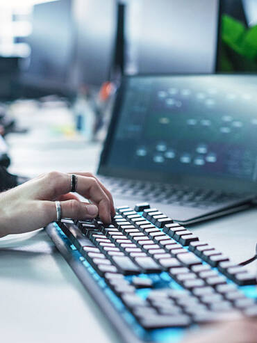 and on mechanical keyboard at computer workstation with multiple monitors showing code and data, headphones nearby, blurred background.