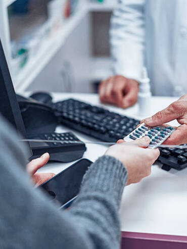 Pharmacist in white coat handing blister pack to patient at pharmacy counter with computer and keyboard visible.