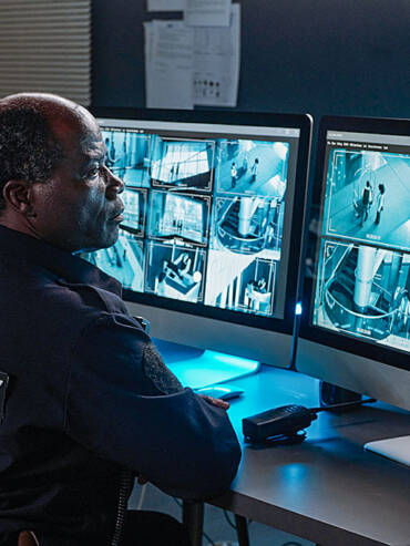 Middle-aged dark-skinned man in security guard uniform intently monitoring multiple security camera feeds on two screens in cool-lit control room.