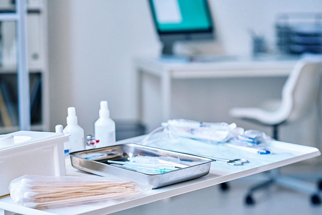 Sterile medical workspace with metal tray holding syringes, cotton swabs, scissors, plastic bottles; computer and chair in background.
