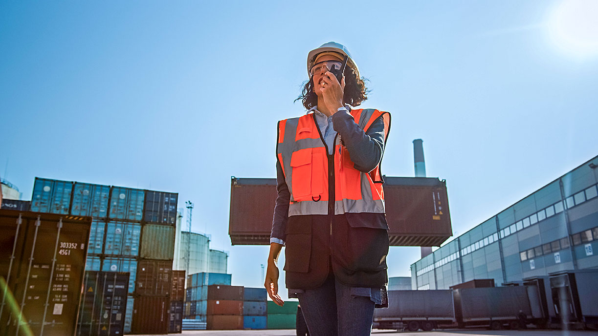 Middle-aged woman in orange safety vest and hard hat talking on radio in sunlit port with shipping containers and cargo.