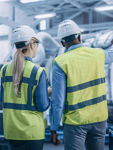 Two workers in light green safety vests and hard hats inspecting equipment in bright, neutral-toned industrial facility.