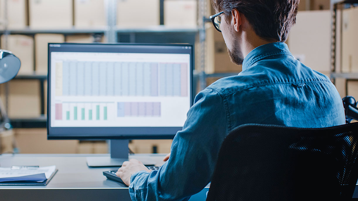 Young male employee in denim shirt focused on dual monitors showing colorful data in warm-lit warehouse with shelves and boxes.
