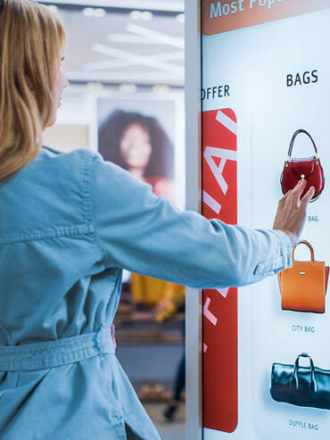 Young woman reaching toward red handbag on digital display in modern retail store with soft lighting and blurred background.