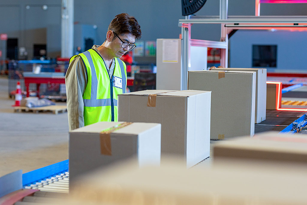 Middle-aged Asian man in yellow safety vest inspects tan cardboard boxes on blue-grey conveyor in bright, efficient warehouse with red scan lights.