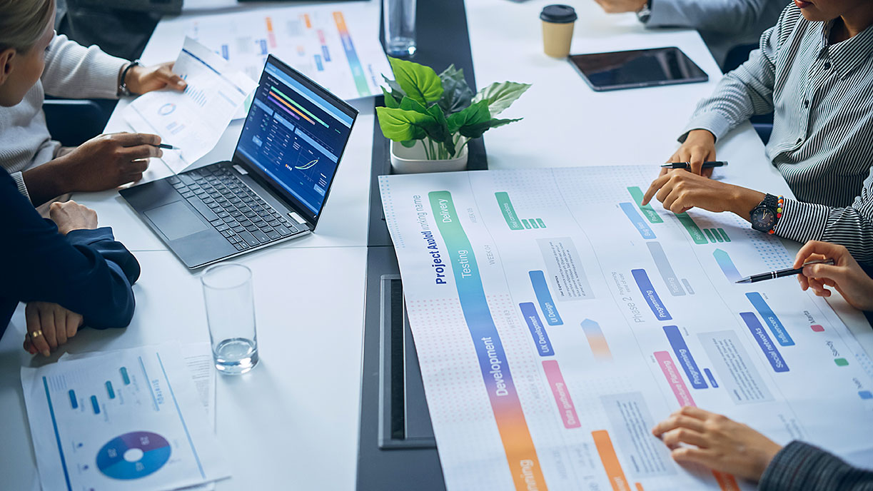 Diverse team collaborates around conference table with laptop showing data analytics, colorful charts, printed documents, and small plant.