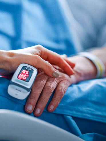 Healthcare professional holding patient’s hand with pulse oximeter showing 96; soft blue hospital linens in background, conveying care.