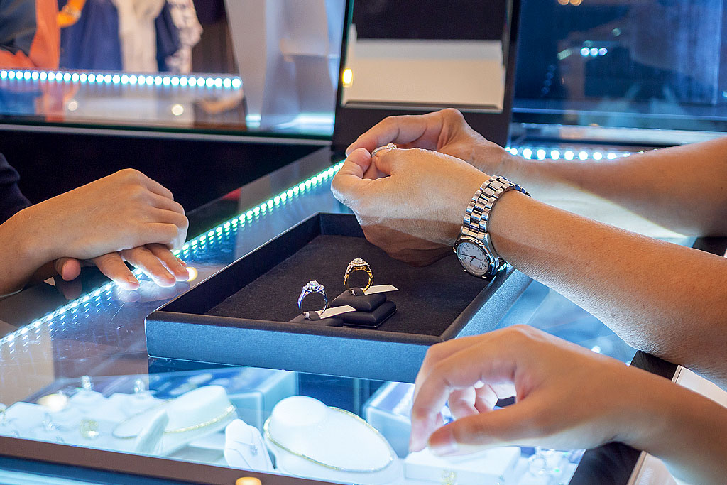 Salesperson holding ring and customer with hand on glass display case, elegant jewelry under soft LED lights in showroom.