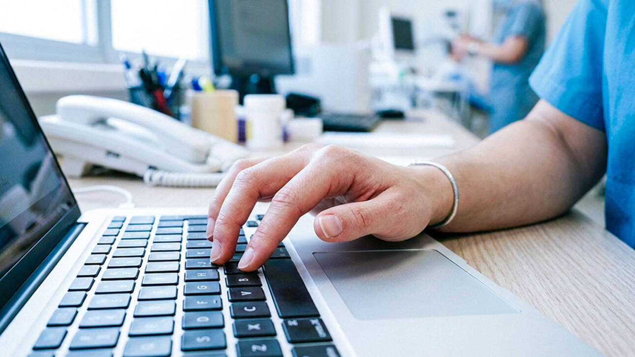Hands in teal uniform typing on a laptop and writing with pen on a document in a softly blurred professional office.