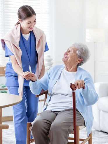 Young caregiver in blue scrubs holding the hand of a smiling elderly woman with cane in a bright sunlit living room.