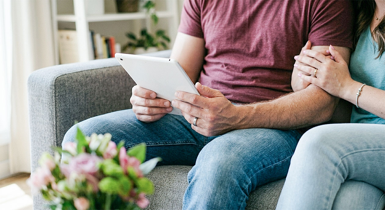 Close-up of young couple's hands holding a white tablet on a gray sofa with soft natural light and blurred flowers.