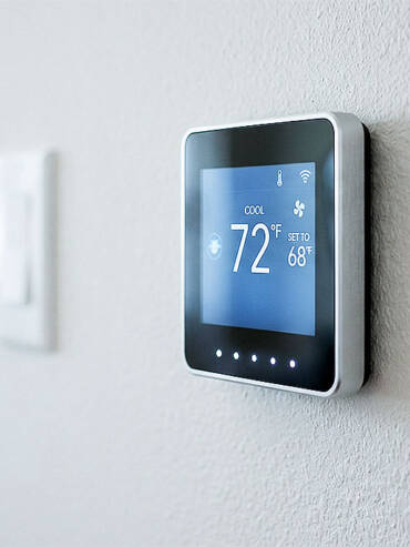 Modern silver-framed digital thermostat with blue screen and white illuminated touch dots on a light beige wall.