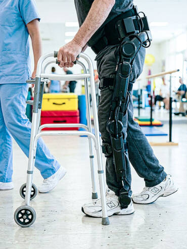 Elderly man in black leg exoskeleton holding silver walker, stepping forward with assistant nearby.