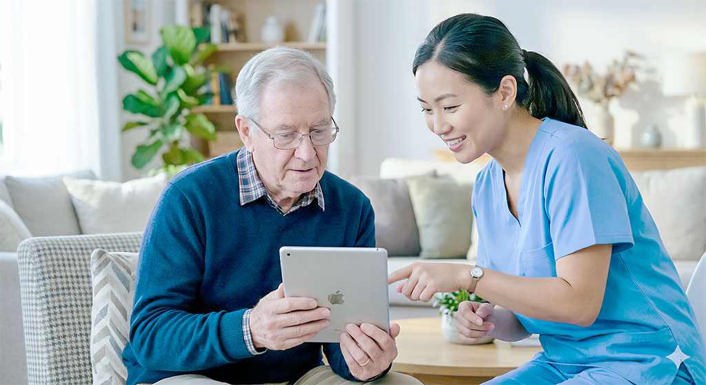 East Asian caregiver in light blue scrubs pointing at tablet held by seated elderly man.
