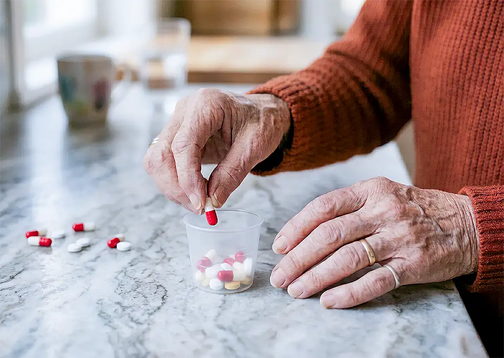 Wrinkled elderly hands sorting pills from a white pill organizer on a countertop.
