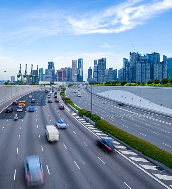 Busy urban highway curves with blurred cars; modern city skyline with glass buildings, cranes, and greenery under partly cloudy sky.