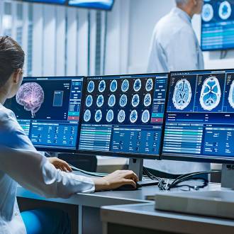 Female scientist in lab coat analyzing brain scans on multiple monitors; two colleagues discuss in background of modern medical lab.